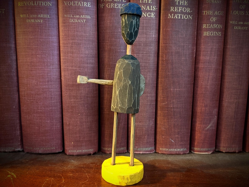 May include: A carved wooden figure with a dark gray body, a blue hat, and a yellow base. The figure has outstretched arms and is positioned in front of a row of vintage books with maroon covers.