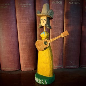 Op de afbeelding: Handgesneden houten beeldje van een persoon die gitaar speelt. De figuur draagt een cowboyhoed en een gele jurk. De basis is groen met de naam "SIERRA" erop geschilderd. De achtergrond toont een rij vintage boeken.