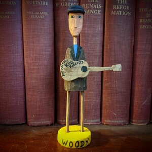 May include: Wooden folk art figure of a man with a guitar. The figure is wearing a cap and jacket, and the guitar has the text "THIS MACHINE KILLS FASCISTS." The base is yellow and says "WOODY."
