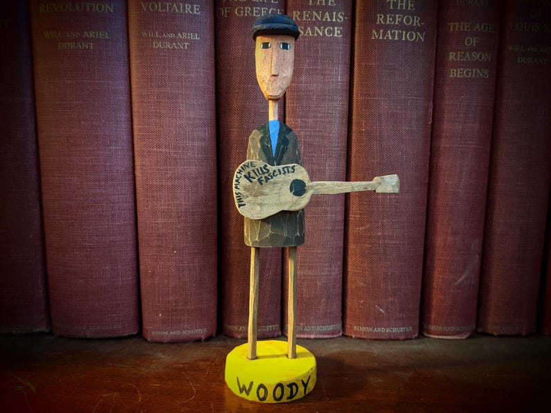 May include: Wooden folk art figure of a man with a guitar. The figure is wearing a cap and jacket, and the guitar has the text "THIS MACHINE KILLS FASCISTS." The base is yellow and says "WOODY."