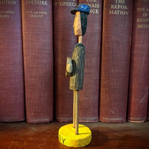 May include: A carved wooden figure of a man wearing a hat and holding a book, standing on a yellow base. The figure is set against a backdrop of antique books with red covers. The base has the word "DY" on it.