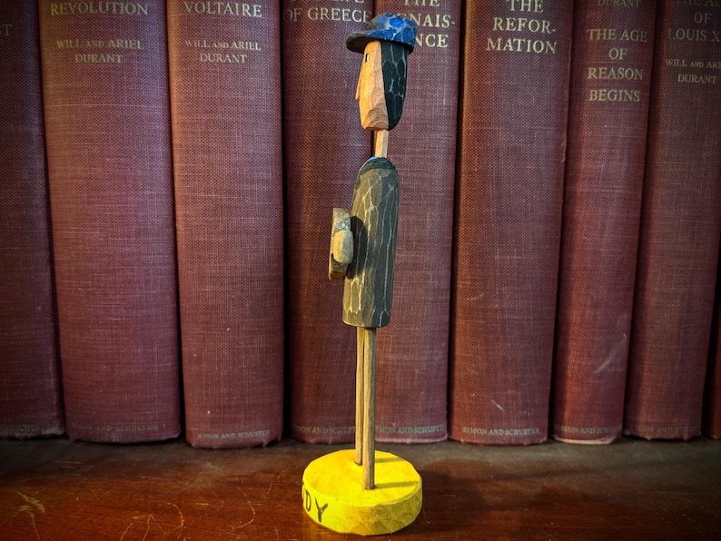 May include: A carved wooden figure of a man wearing a hat and holding a book, standing on a yellow base. The figure is set against a backdrop of antique books with red covers. The base has the word "DY" on it.