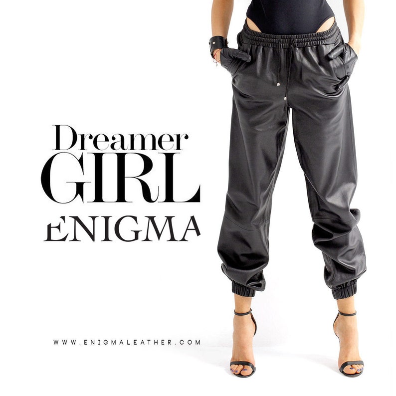 May include: Black leather jogger pants with a drawstring waist and elastic cuffs. The pants are styled with a black top and black heeled sandals. The text "Dreamer Girl Enigma" and "www.enigmaleather.com" are printed below the image.