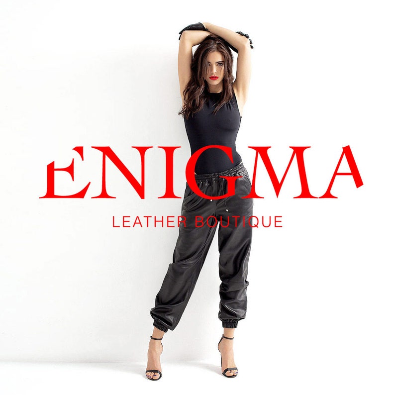 May include: A woman wearing a black bodysuit and black leather pants with a drawstring waist. She is wearing black heeled shoes and black leather gloves. The text "ENIGMA LEATHER BOUTIQUE" is displayed in red letters.