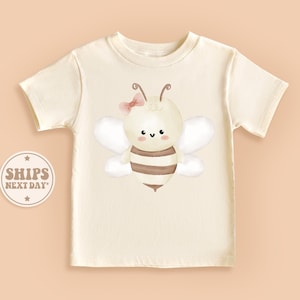 May include: A white t-shirt with a cute cartoon bee wearing a pink bow. The bee has brown and yellow stripes and a smiling face.