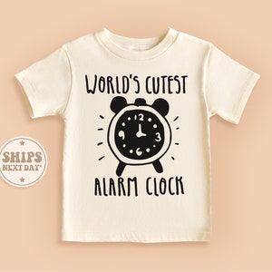 May include: Cream-colored short-sleeved t-shirt with a black graphic of an alarm clock and the text "WORLD'S CUTEST ALARM CLOCK". The design features a classic alarm clock silhouette with radiating lines.