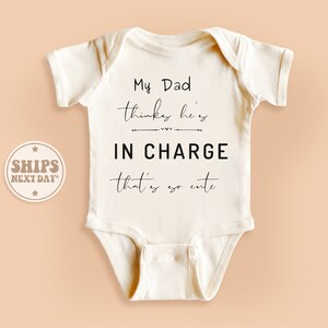 May include: White baby onesie with black text that reads "My Dad thinks he's IN CHARGE that's so cute".