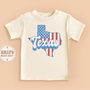 May include: A white t-shirt with a vintage-style graphic of the state of Texas, designed with the American flag colors. The text "Texas" is in a retro font.