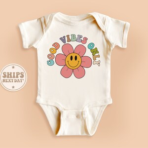 May include: A white baby onesie with a pink flower and the text "GOOD VIBES ONLY" in rainbow colors.