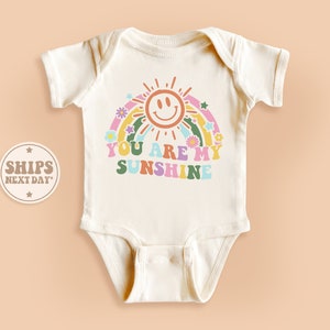 May include: A white baby onesie with a rainbow, sun, and the text "You are my sunshine" in colorful retro lettering.