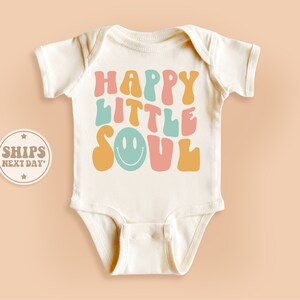 May include: A white baby onesie with the text "HAPPY LITTLE SOUL" in colorful, retro-style lettering. A smiley face is included in the design.