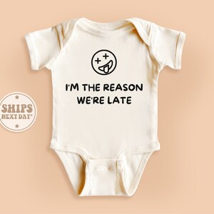 May include: Cream-colored baby onesie with the text "I'M THE REASON WE'RE LATE" in black. Above the text is a black smiley face with crossed-out eyes and a tongue sticking out. The onesie is a short-sleeved garment.