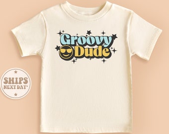 May include: A white t-shirt with a retro-style graphic that says "Groovy Dude" in blue, yellow, and black with a smiling face wearing sunglasses.