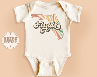 May include: A white baby onesie with a vintage-style graphic of the state of Florida. The graphic features a colorful sunburst design and the word "Florida" in a retro font.