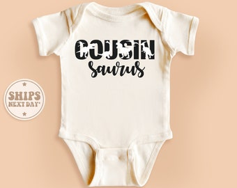 May include: Cream-colored baby onesie with the words "COUSIN saurus" in black script and dinosaur silhouettes. The onesie has short sleeves and a round neckline. The text is centered on the front of the garment.
