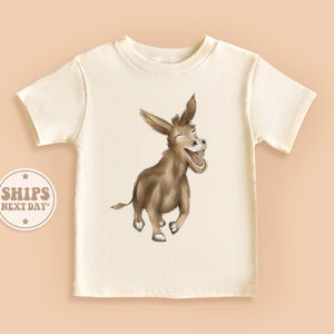 May include: A white t-shirt with a brown donkey illustration. The donkey is smiling and has its mouth open. The donkey is walking and has its legs in the air.