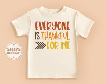 May include: Cream-colored short-sleeved t-shirt with the text "EVERYONE IS THANKFUL FOR ME" in shades of orange, brown, and yellow. A series of arrows point to the right. A round sticker with the words "SHIPS NEXT DAY" is on the left.