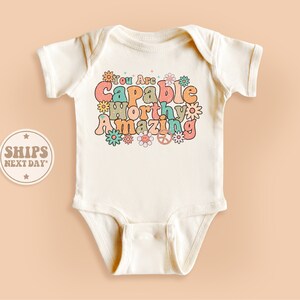 May include: White baby onesie with a colorful retro-style graphic that says "You Are Capable Worthy Amazing" with flowers and peace signs.