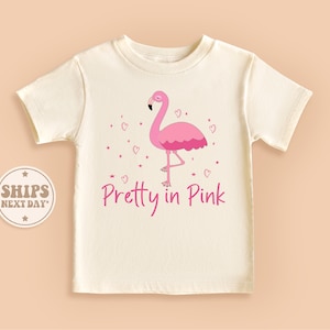 May include: A white t-shirt with a pink flamingo and pink hearts. The text "Pretty in Pink" is printed below the flamingo.
