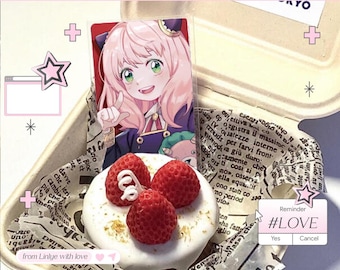 May include: Anya Forger character acrylic standee with a white frosted donut topped with three strawberries. The donut is in a takeout container with printed paper. A reminder with the hashtag #LOVE is also visible.