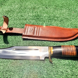 Puede incluir: Un cuchillo de hoja fija con una hoja de acero pulido, una guarda de latón y un mango con anillos apilados en negro y marrón. El cuchillo viene acompañado de una funda de cuero marrón con cierre de broche y un logotipo grabado en el frente.