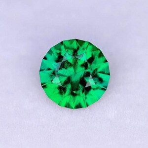 May include: A green gemstone with a faceted cut. The gemstone has a dark green color with black inclusions.