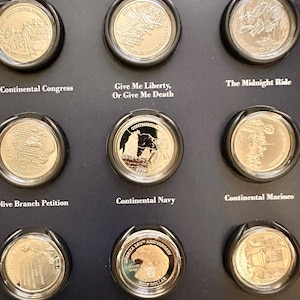 Puede incluir: Una colección de nueve monedas conmemorativas de tono plateado exhibidas en un soporte negro. Cada moneda presenta imágenes históricas únicas y etiquetas de texto que incluyen Continental Congress, Give Me Liberty, Or Give Me Death, The Midnight Ride, Olive Branch Petition, Continental Navy, Continental Marines, Lee Resolution, Adopting Independence y Proclaim Liberty.