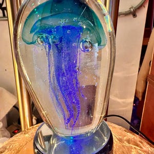 以下が含まれることがあります： 青とティールアクセントの装飾的なガラスクラゲ彫刻。クラゲは透明なガラスに囲まれ、ベースを照らす青い光があります。彫刻は丸いベースと球根状のトップを備え、ユニークで芸術的なデザインを作り出しています。