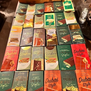 May include: A large collection of rectangular chocolate bars arranged in rows, featuring various brands and packaging designs labelled Dubai Style. The bars display illustrations of chocolate with pistachio and kadaif fillings, with net weights of 200g.
