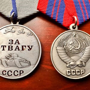 Può includere: Due medaglie d'argento con nastri rossi e blu. La medaglia a sinistra presenta un carro armato e il testo "ЗА ОТВАГУ" e "CCCP". La medaglia a destra presenta un globo alato con una falce e un martello e il testo "CCCP".