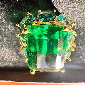 May include: A large emerald green gemstone ring with a gold setting. The ring is surrounded by smaller, light blue gemstones.