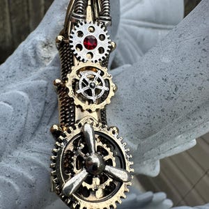 Peut inclure: Une pièce d'armure de doigt en métal doré avec une base texturée ornée de trois engrenages argentés superposés. L'engrenage supérieur inclut un petit strass rouge, celui du milieu a un motif de roue de navire et l'inférieur une hélice.