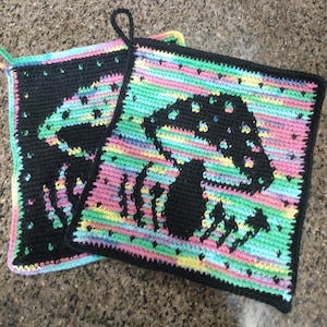 May include: Two crocheted pot holders with a mushroom design. One pot holder is black with a white mushroom design, and the other is multi-colored with a black mushroom design.