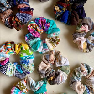 Puede incluir: Una colección de coloridas gomas para el pelo de tela con varios patrones y colores, incluyendo diseños florales, a rayas y patchwork. Las gomas para el pelo están dispuestas sobre una superficie de color claro, mostrando su calidad artesanal y estética diversa.