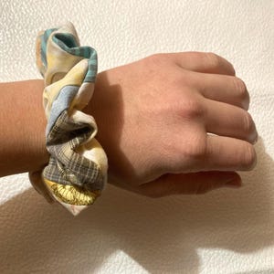 K&ouml;nnte beinhalten: Ein Haargummi mit Patchwork-Design. Der Stoff weist eine Mischung aus Mustern auf, darunter Karos, Blumenmuster und unifarbene Farben in Gelb-, Blau- und Braunt&ouml;nen. Das Haargummi ist um ein Handgelenk gewickelt.