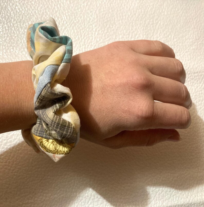 K&ouml;nnte beinhalten: Ein Haargummi mit Patchwork-Design. Der Stoff weist eine Mischung aus Mustern auf, darunter Karos, Blumenmuster und unifarbene Farben in Gelb-, Blau- und Braunt&ouml;nen. Das Haargummi ist um ein Handgelenk gewickelt.