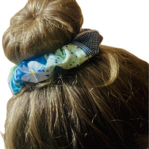 K&ouml;nnte beinhalten: Ein farbenfrohes Haargummi mit Blumen- und Tupfenmuster umwickelt einen Dutt aus braunem Haar. Das Haargummi hat einen Stoff mit blauen, wei&szlig;en und gelben Blumen sowie einen Abschnitt aus schwarz-wei&szlig; kariertem Stoff.