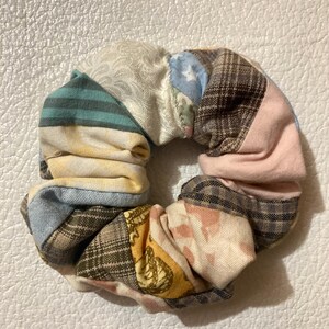 K&ouml;nnte beinhalten: Ein farbenfrohes Scrunchie aus verschiedenen Stoffresten. Das Scrunchie hat ein Patchwork-Design mit Streifen, Karos und floralen Mustern in Blau-, Rosa-, Gelb- und Braunt&ouml;nen. Ein stilvolles Haaraccessoire f&uuml;r jeden Look.