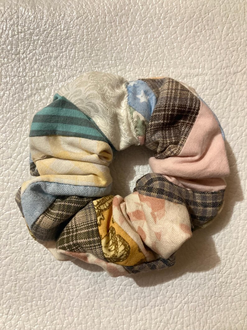 K&ouml;nnte beinhalten: Ein farbenfrohes Scrunchie aus verschiedenen Stoffresten. Das Scrunchie hat ein Patchwork-Design mit Streifen, Karos und floralen Mustern in Blau-, Rosa-, Gelb- und Braunt&ouml;nen. Ein stilvolles Haaraccessoire f&uuml;r jeden Look.