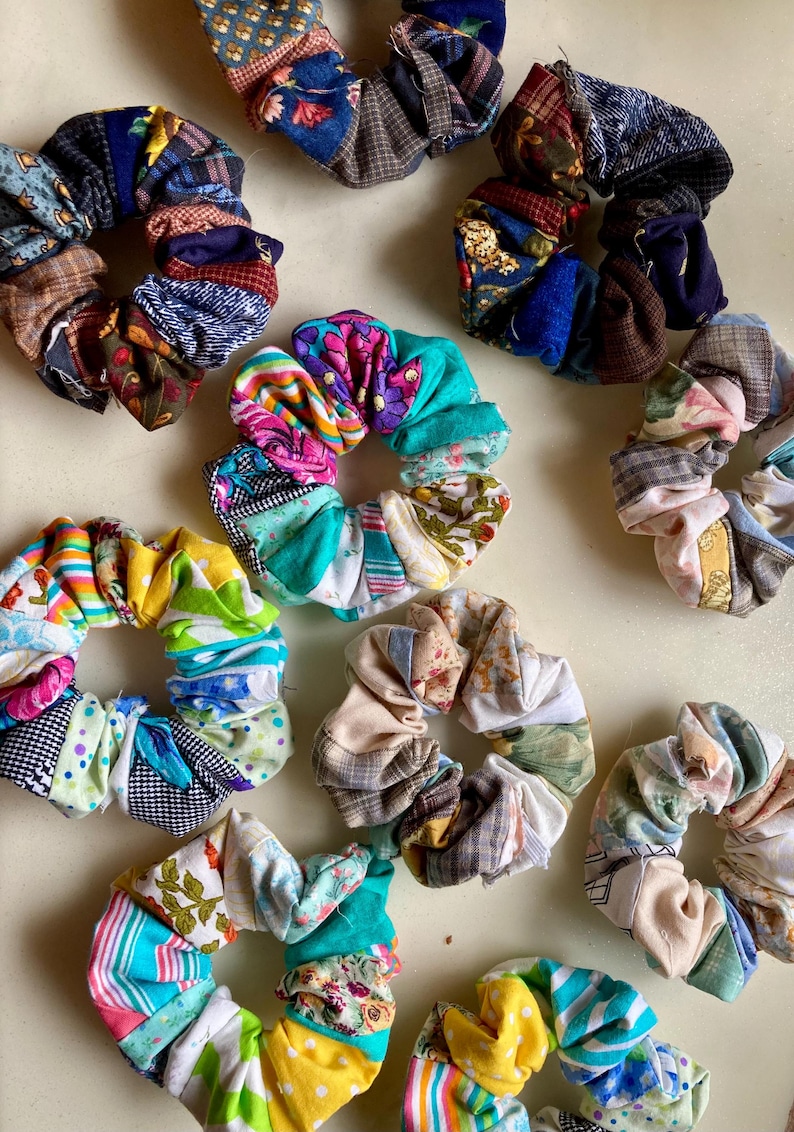 K&ouml;nnte beinhalten: Eine Sammlung bunter Stoff-Haargummis in verschiedenen Mustern und Farben, darunter Blumen-, Streifen- und Patchwork-Designs. Die Haargummis sind auf einer hellen Oberfl&auml;che angeordnet und zeigen ihre handgefertigte Qualit&auml;t und vielf&auml;ltige &Auml;sthetik.