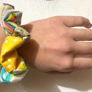 K&ouml;nnte beinhalten: Ein mehrfarbiges Scrunchie mit Patchwork-Design. Das Scrunchie hat eine Vielzahl von Stoffen, darunter Gelb mit wei&szlig;en Punkten, Streifenmuster und Blumendrucke. Das Scrunchie ist um ein Handgelenk gewickelt.