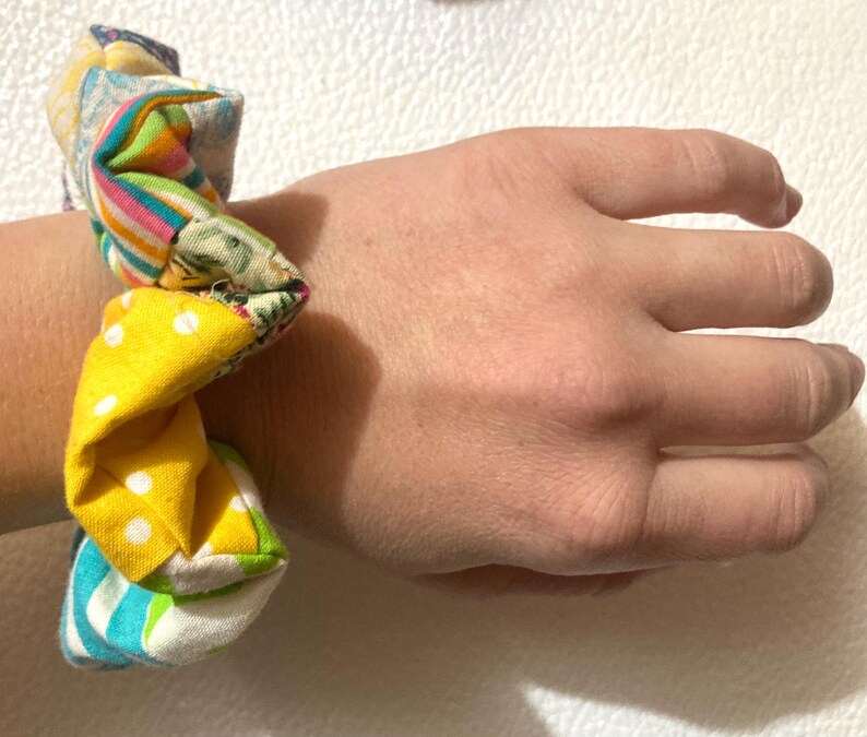K&ouml;nnte beinhalten: Ein mehrfarbiges Scrunchie mit Patchwork-Design. Das Scrunchie hat eine Vielzahl von Stoffen, darunter Gelb mit wei&szlig;en Punkten, Streifenmuster und Blumendrucke. Das Scrunchie ist um ein Handgelenk gewickelt.