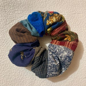 K&ouml;nnte beinhalten: Ein farbenfrohes Scrunchie aus verschiedenen Stoffresten. Das Scrunchie hat ein Patchwork-Design mit Blau-, Braun-, Rot- und Gr&uuml;nt&ouml;nen. Die Stoffmuster umfassen florale, geometrische und strukturierte Designs. Dieses Haaraccessoire ist eine einzigartige und stilvolle Erg&auml;nzung.