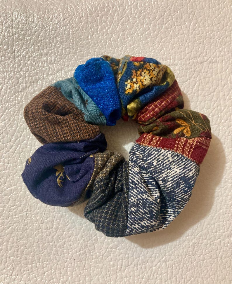K&ouml;nnte beinhalten: Ein farbenfrohes Scrunchie aus verschiedenen Stoffresten. Das Scrunchie hat ein Patchwork-Design mit Blau-, Braun-, Rot- und Gr&uuml;nt&ouml;nen. Die Stoffmuster umfassen florale, geometrische und strukturierte Designs. Dieses Haaraccessoire ist eine einzigartige und stilvolle Erg&auml;nzung.