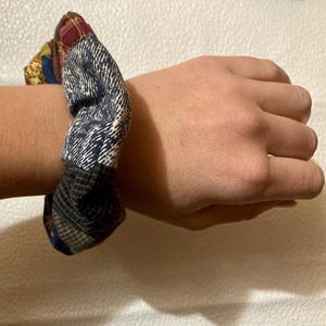 K&ouml;nnte beinhalten: Ein Patchwork-Haargummi in Blau-, Grau-, Rot- und Goldt&ouml;nen ist um ein Handgelenk gewickelt. Der Stoff weist verschiedene Muster auf, darunter Karos und Fischgr&auml;ten. Dieses Haaraccessoire verleiht einen Vintage-Touch.