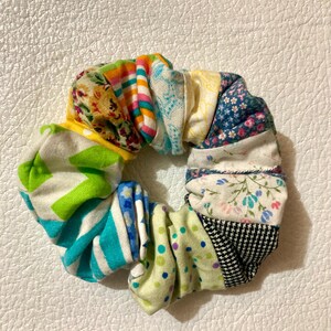 K&ouml;nnte beinhalten: Ein mehrfarbiges Haargummi aus Stoffresten. Das Haargummi hat ein Patchwork-Design mit floralen, gestreiften und gepunkteten Mustern in Gelb-, Gr&uuml;n-, Blau- und Rosat&ouml;nen. Das Haaraccessoire ist kreisf&ouml;rmig und scheint handgefertigt zu sein.