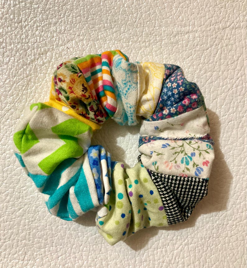 K&ouml;nnte beinhalten: Ein mehrfarbiges Haargummi aus Stoffresten. Das Haargummi hat ein Patchwork-Design mit floralen, gestreiften und gepunkteten Mustern in Gelb-, Gr&uuml;n-, Blau- und Rosat&ouml;nen. Das Haaraccessoire ist kreisf&ouml;rmig und scheint handgefertigt zu sein.