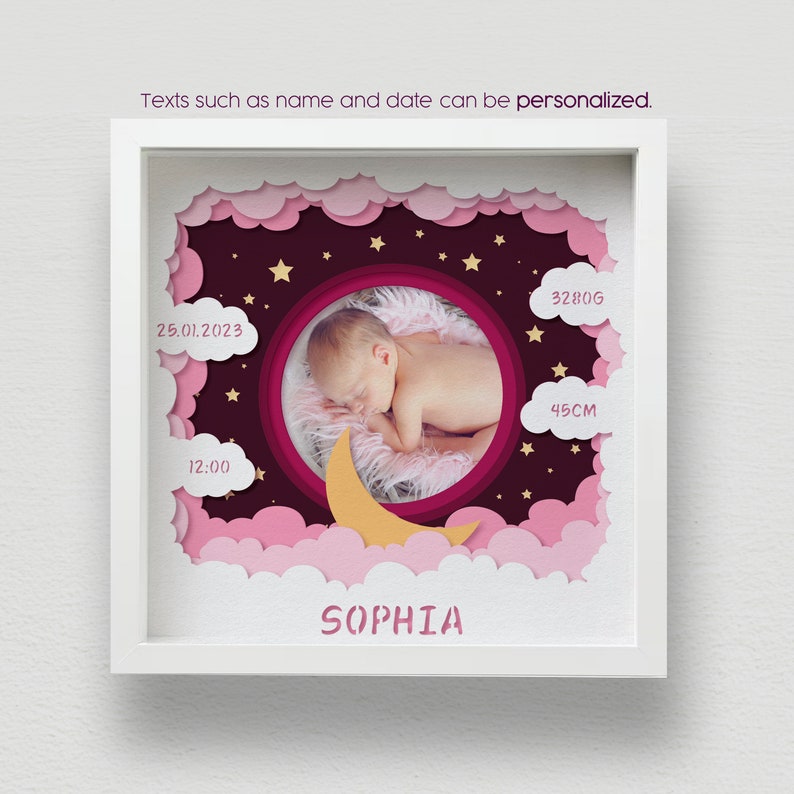 May include: A framed, layered paper art piece featuring a sleeping baby in a circular frame. The design includes pink clouds, stars, and a crescent moon. Text includes the baby's name, birth date, time, weight (3280g), and length (45cm).