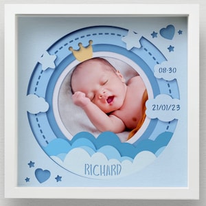 May include: A framed baby photo with a blue and white layered design. The photo features a sleeping infant wearing a small gold crown. The frame includes the baby's name, birth time, and date.