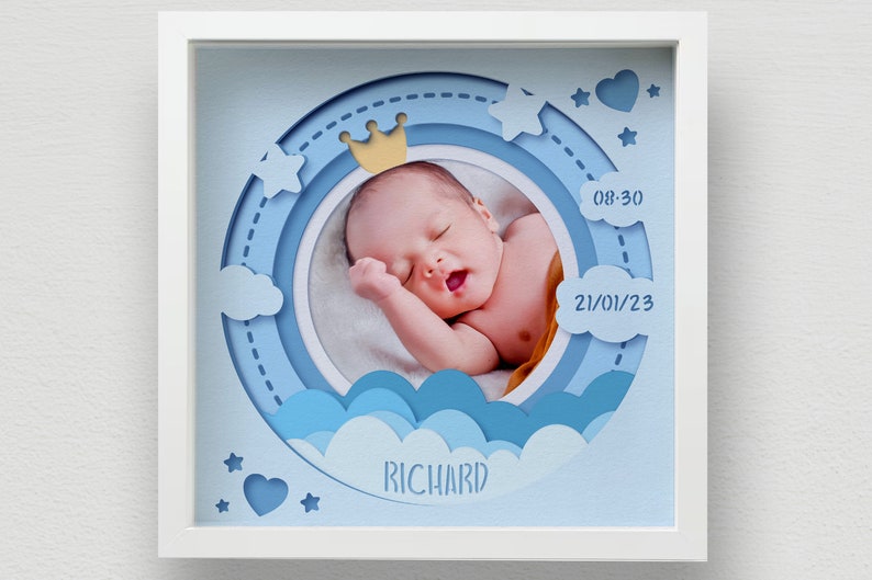 May include: A framed baby photo with a blue and white layered design. The photo features a sleeping infant wearing a small gold crown. The frame includes the baby's name, birth time, and date.